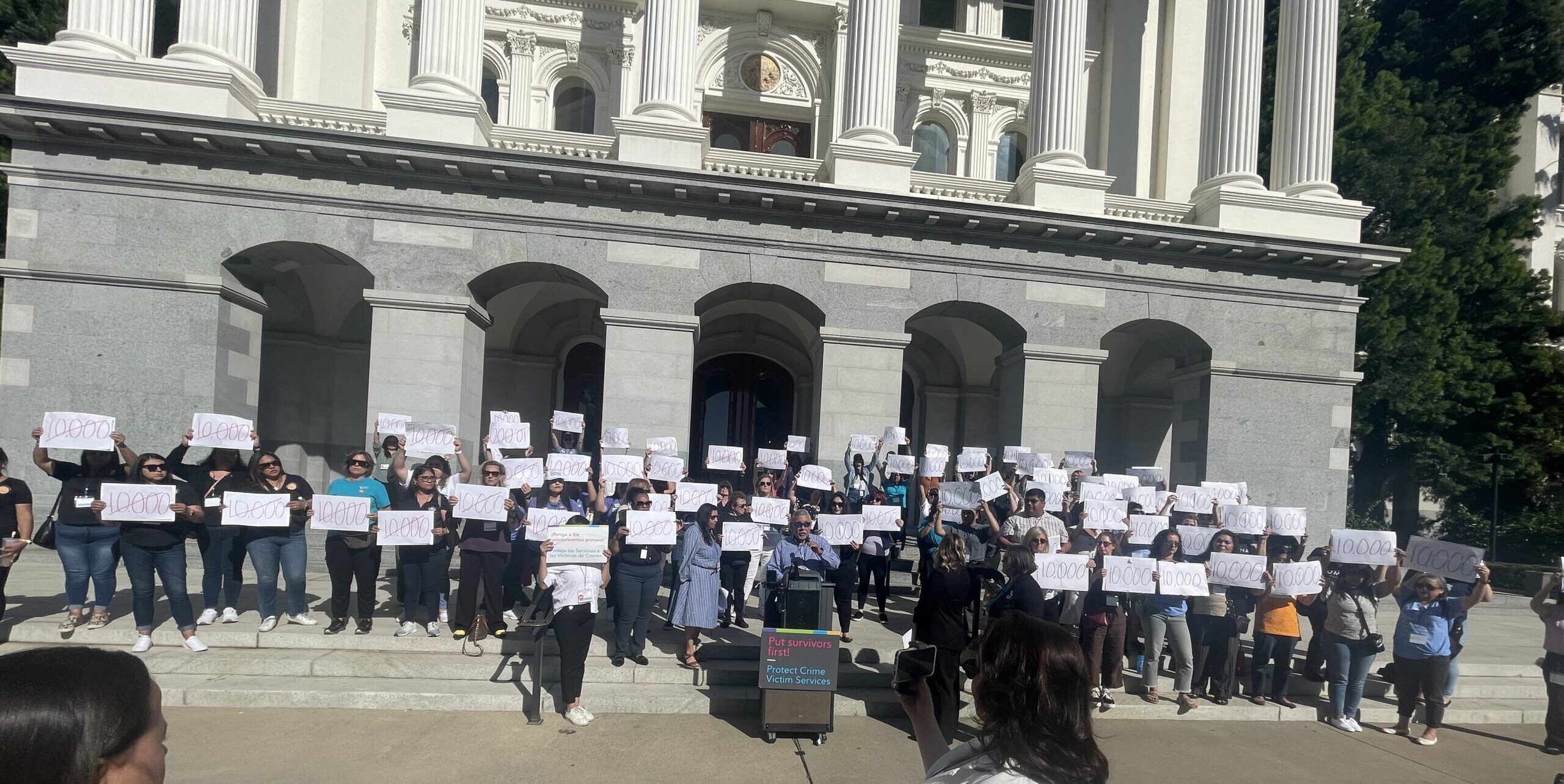 Photo of VALOR and the California Partnership to End Domestic Violence Rally. Participants are standing on the West Steps of the California Capitol holdings signs that show the number 10,000 crossed out in red ink.