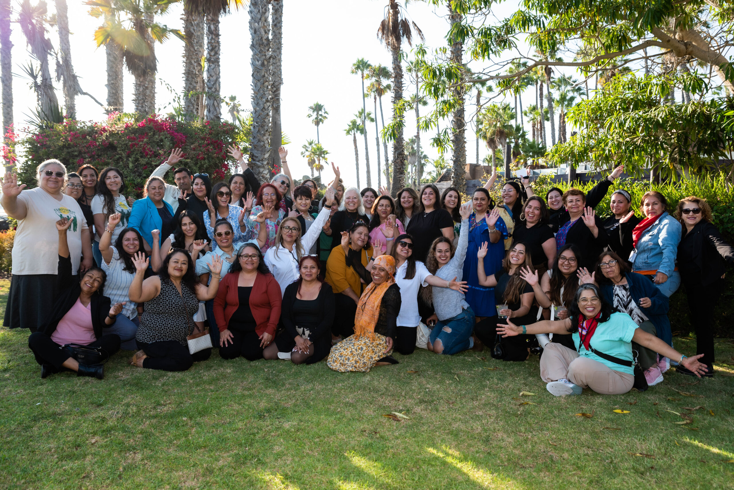 Group photo in front of palm trees and bushes with some people sitting and kneeling on grass.