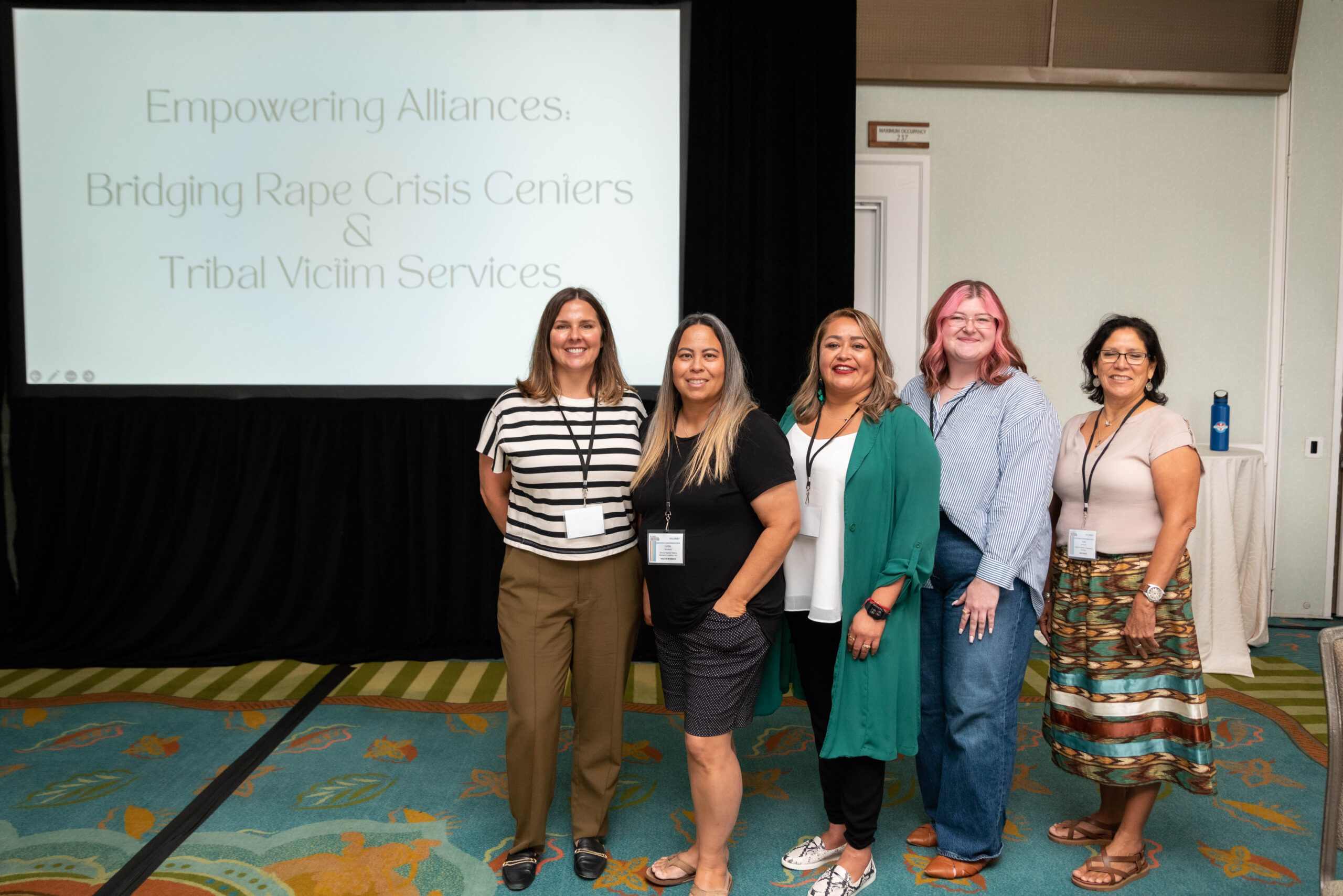 5 people stand in a room in front of a projector screen that has the text "Empowering Alliances: Bridging Rape Crisis Centers & Tribal Victim Services."